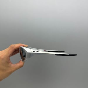 Pu&ograve; includere: Occhiali da sole argentati con lenti scure. Gli occhiali hanno un design elegante con aste nere e un piccolo dettaglio rosso. Sono tenuti da una mano su sfondo grigio.