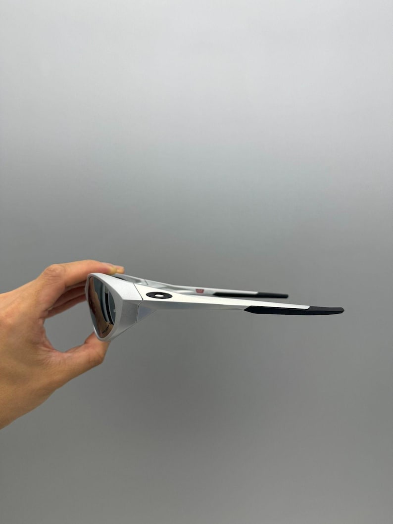 Pu&ograve; includere: Occhiali da sole argentati con lenti scure. Gli occhiali hanno un design elegante con aste nere e un piccolo dettaglio rosso. Sono tenuti da una mano su sfondo grigio.