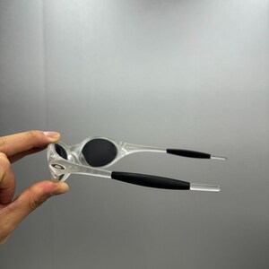 May include: Clear-framed sunglasses with black lenses and black arm grips. The sunglasses have a sporty design and are held up in the air. The Oakley logo is visible on the frame.