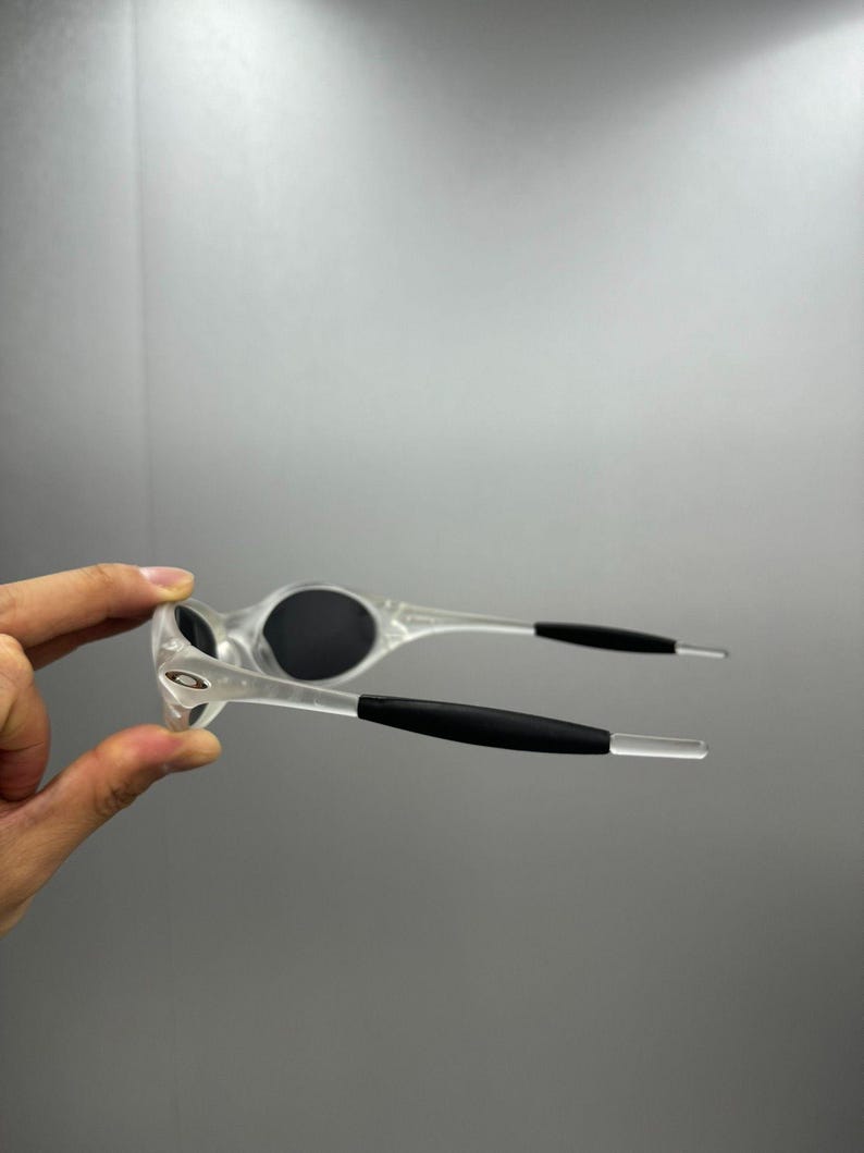 May include: Clear-framed sunglasses with black lenses and black arm grips. The sunglasses have a sporty design and are held up in the air. The Oakley logo is visible on the frame.