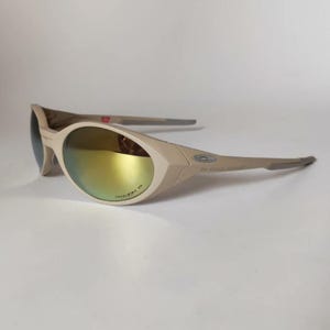 Puede incluir: Gafas de sol de color beige con un diseño de montura envolvente y lentes reflectantes con tonos dorados. Las patillas muestran un emblema de logotipo metálico y la superficie de la lente tiene grabado el texto Prizm P cerca del borde inferior.
