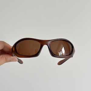 May include: Brown sunglasses with a dark frame and tinted lenses. The sunglasses have a sporty, wraparound design. The lenses are a medium brown colour, and the frame is a darker shade of brown.