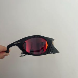 Może przedstawiać: Czarne okulary przeciwsłoneczne z czerwonymi soczewkami. Okulary mają elegancki, nowoczesny design z zakrzywioną oprawką i małym logo na zauszniku. Soczewki są w żywym czerwonym kolorze.