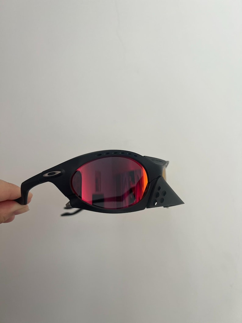 Może przedstawiać: Czarne okulary przeciwsłoneczne z czerwonymi soczewkami. Okulary mają elegancki, nowoczesny design z zakrzywioną oprawką i małym logo na zauszniku. Soczewki są w żywym czerwonym kolorze.