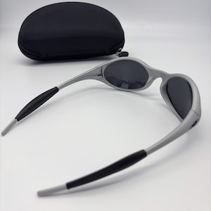 Puede incluir: Gafas de sol grises con lentes oscuras y puntas de patillas negras. Las gafas de sol tienen un dise&ntilde;o elegante y moderno. Un estuche negro de forma ovalada est&aacute; en el fondo.