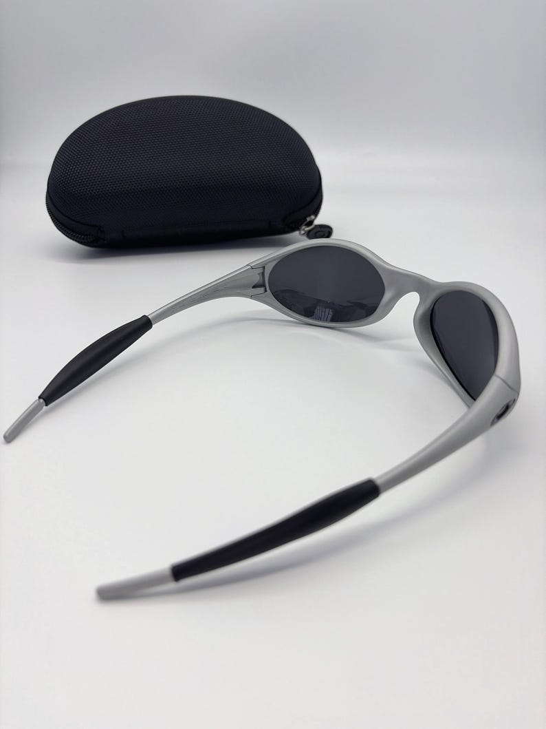 Puede incluir: Gafas de sol grises con lentes oscuras y puntas de patillas negras. Las gafas de sol tienen un dise&ntilde;o elegante y moderno. Un estuche negro de forma ovalada est&aacute; en el fondo.