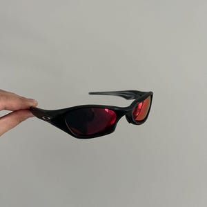 May include: Black sunglasses with red lenses. The sunglasses have a sleek, curved design and a matte finish. The lenses are a vibrant red colour, contrasting with the black frames. The sunglasses are held against a plain background.