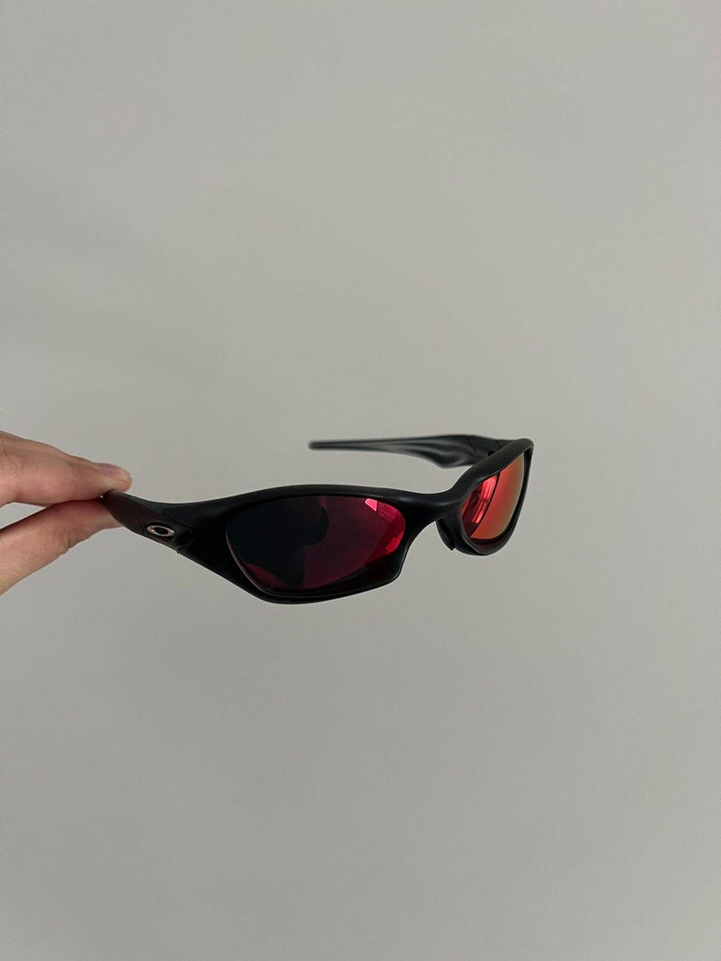 May include: Black sunglasses with red lenses. The sunglasses have a sleek, curved design and a matte finish. The lenses are a vibrant red colour, contrasting with the black frames. The sunglasses are held against a plain background.