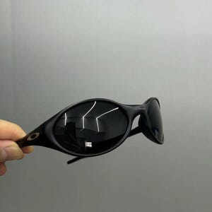 Puede incluir: Gafas de sol negras de forma ovalada con lentes oscuras. Las gafas tienen un dise&ntilde;o elegante y moderno con una montura curvada y un peque&ntilde;o logotipo en la patilla. Las lentes reflejan la luz.
