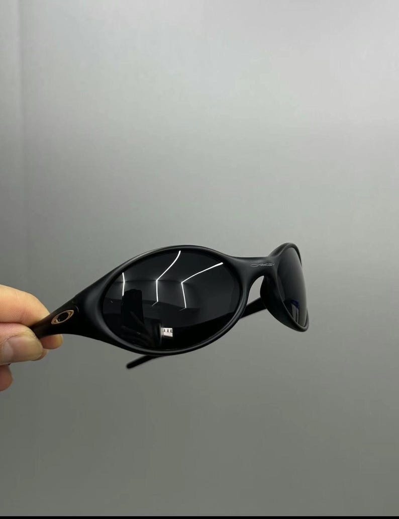Puede incluir: Gafas de sol negras de forma ovalada con lentes oscuras. Las gafas tienen un dise&ntilde;o elegante y moderno con una montura curvada y un peque&ntilde;o logotipo en la patilla. Las lentes reflejan la luz.