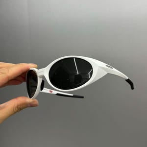 K&ouml;nnte beinhalten: Wei&szlig;e Sonnenbrille mit gro&szlig;en, ovalen Gl&auml;sern und einem schlanken Design. Die Rahmen sind wei&szlig; mit schwarzen Akzenten an den B&uuml;geln. Die dunklen Gl&auml;ser bilden einen Kontrast zu den wei&szlig;en Rahmen. Ein stilvolles Accessoire zum Schutz der Augen.