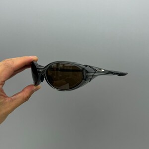 May include: Black sunglasses with brown lenses. The sunglasses have a wraparound design with a thick frame and a small Oakley logo on the temple. The lenses are tinted, and the frame is a glossy black.