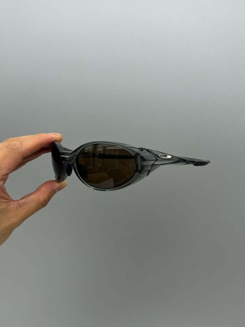 May include: Black sunglasses with brown lenses. The sunglasses have a wraparound design with a thick frame and a small Oakley logo on the temple. The lenses are tinted, and the frame is a glossy black.