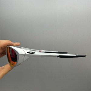 Puede incluir: Gafas de sol blancas con lentes rojo-naranja. Las monturas son blancas con detalles negros en las patillas. Las lentes tienen un revestimiento reflectante. Las gafas de sol est&aacute;n sujetas por una mano sobre un fondo gris.
