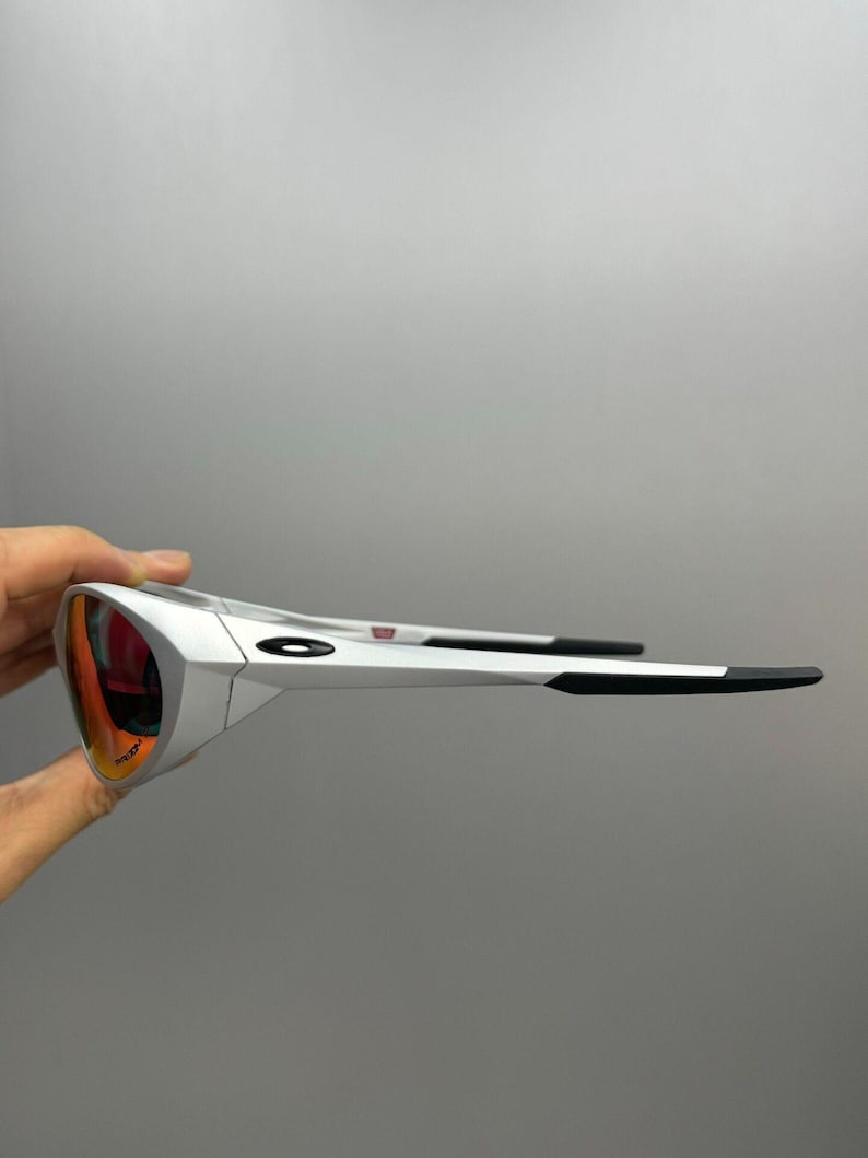 Puede incluir: Gafas de sol blancas con lentes rojo-naranja. Las monturas son blancas con detalles negros en las patillas. Las lentes tienen un revestimiento reflectante. Las gafas de sol est&aacute;n sujetas por una mano sobre un fondo gris.