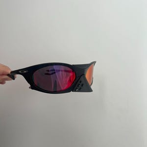 Może przedstawiać: Czarne okulary przeciwsłoneczne z czerwonymi i pomarańczowymi soczewkami. Okulary mają elegancki, nowoczesny design z zakrzywioną oprawką i małym, tr&oacute;jkątnym detalem w pobliżu soczewek. Soczewki odbijają światło.
