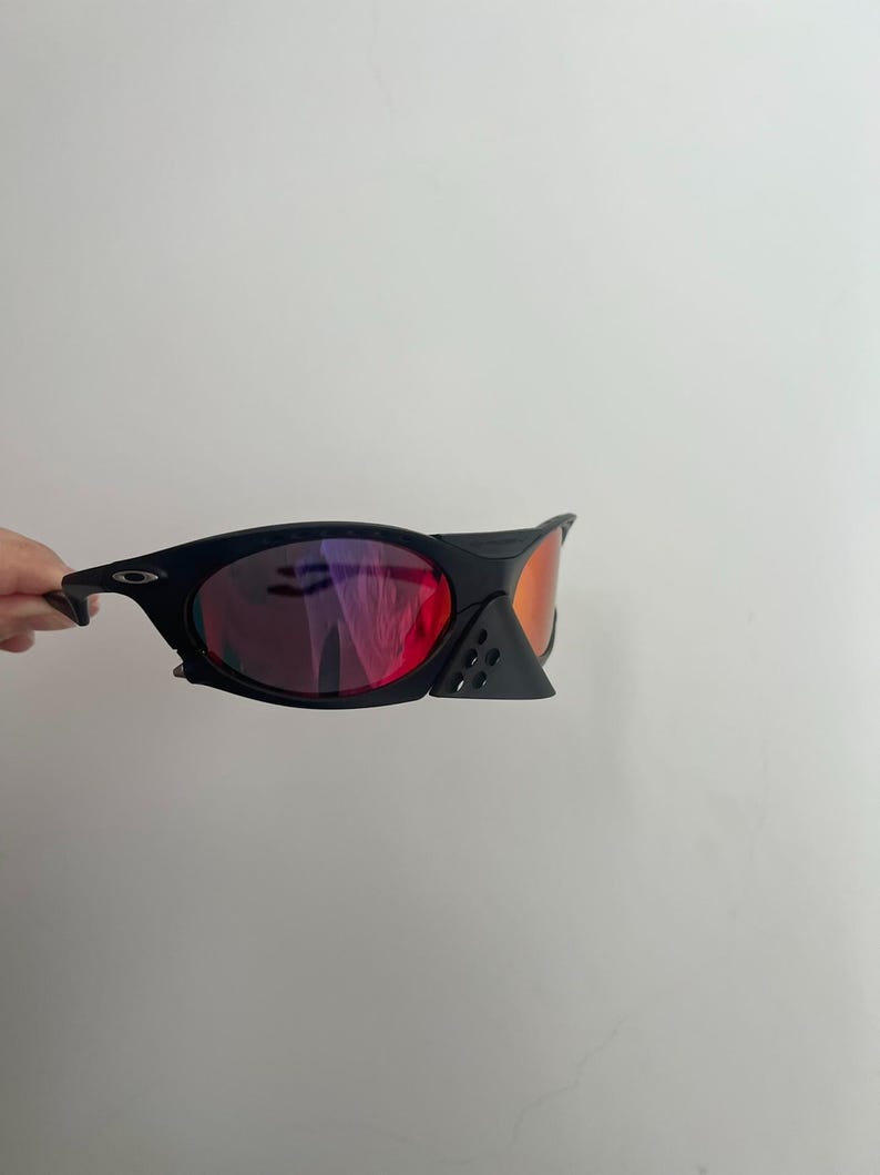 Może przedstawiać: Czarne okulary przeciwsłoneczne z czerwonymi i pomarańczowymi soczewkami. Okulary mają elegancki, nowoczesny design z zakrzywioną oprawką i małym, tr&oacute;jkątnym detalem w pobliżu soczewek. Soczewki odbijają światło.