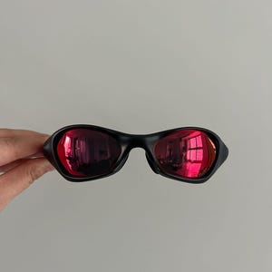 May include: Black sunglasses with red mirrored lenses. The sunglasses have a sleek, modern design with a curved frame. The lenses reflect the surrounding environment, creating a vibrant effect. The sunglasses are held by a hand.