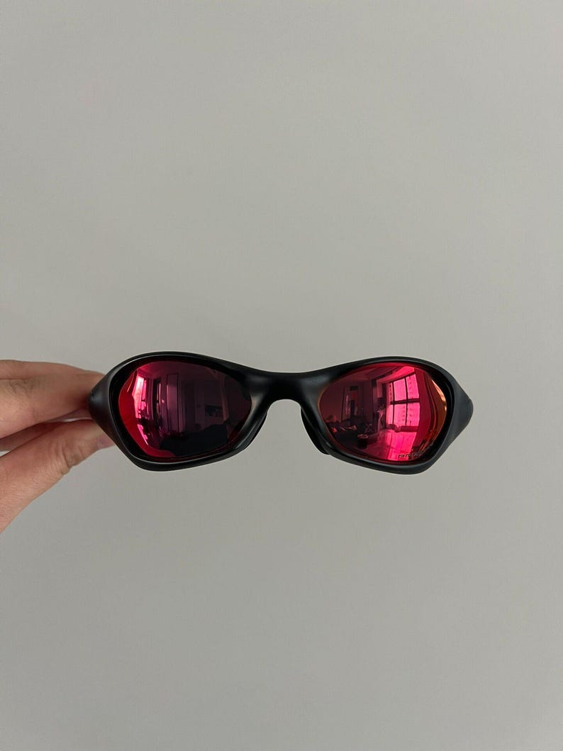 May include: Black sunglasses with red mirrored lenses. The sunglasses have a sleek, modern design with a curved frame. The lenses reflect the surrounding environment, creating a vibrant effect. The sunglasses are held by a hand.