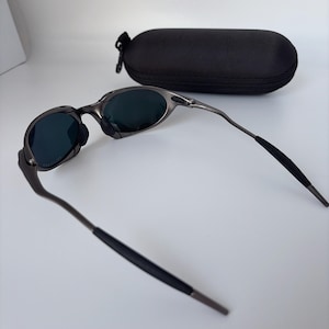 May include: Gunmetal sunglasses with dark lenses and a black protective case. The sunglasses feature a modern design with curved arms and a nose bridge. The oval-shaped case has a zip closure, offering secure storage for the eyewear.