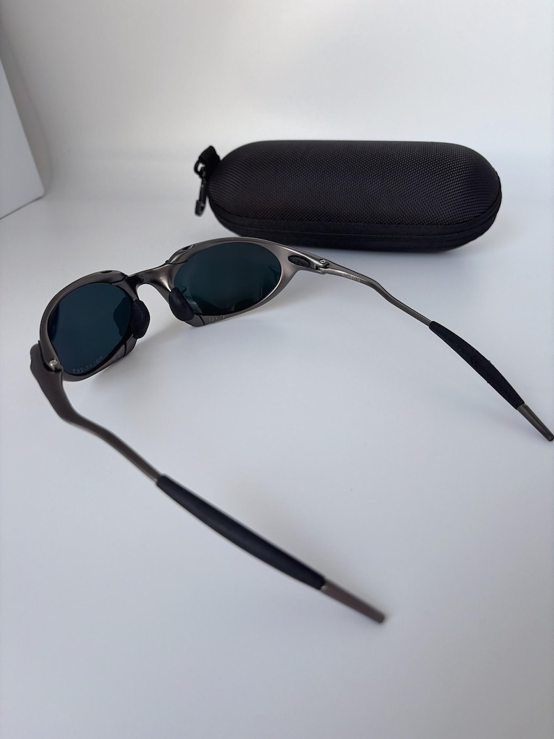 May include: Gunmetal sunglasses with dark lenses and a black protective case. The sunglasses feature a modern design with curved arms and a nose bridge. The oval-shaped case has a zip closure, offering secure storage for the eyewear.