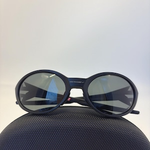 May include: Black sunglasses with round lenses and a thick frame, resting on a textured black case. The lenses reflect the surrounding environment. The brand name "Oakley" is visible on the frame.