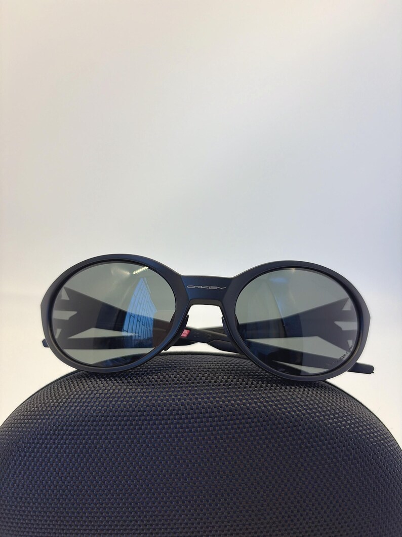 May include: Black sunglasses with round lenses and a thick frame, resting on a textured black case. The lenses reflect the surrounding environment. The brand name "Oakley" is visible on the frame.
