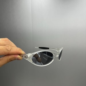 May include: Clear-framed sunglasses with dark lenses. The sunglasses have a sleek, modern design with a curved frame and a black temple arm. The lenses are oval-shaped and reflect the surrounding environment.
