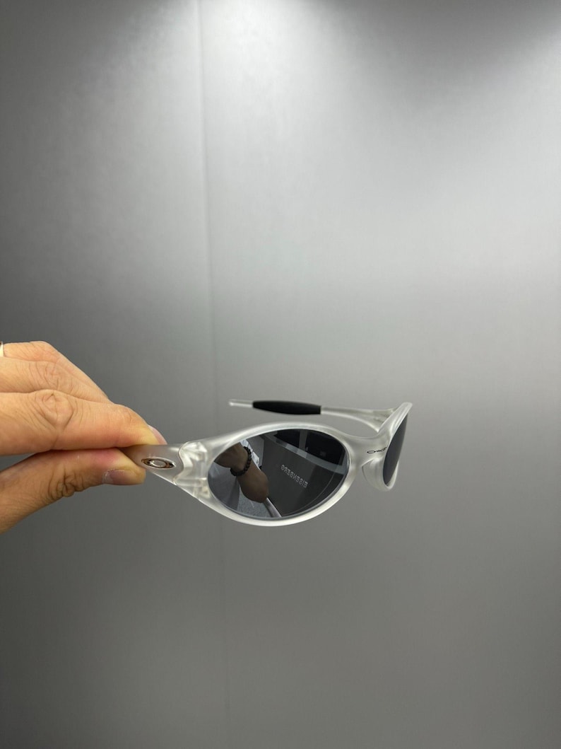 May include: Clear-framed sunglasses with dark lenses. The sunglasses have a sleek, modern design with a curved frame and a black temple arm. The lenses are oval-shaped and reflect the surrounding environment.
