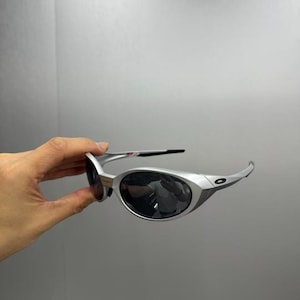 Pode incluir: &Oacute;culos de sol prateados com lentes escuras. Os &oacute;culos t&ecirc;m um design moderno com hastes curvas e uma forma de lente arredondada. As lentes escuras protegem do sol.