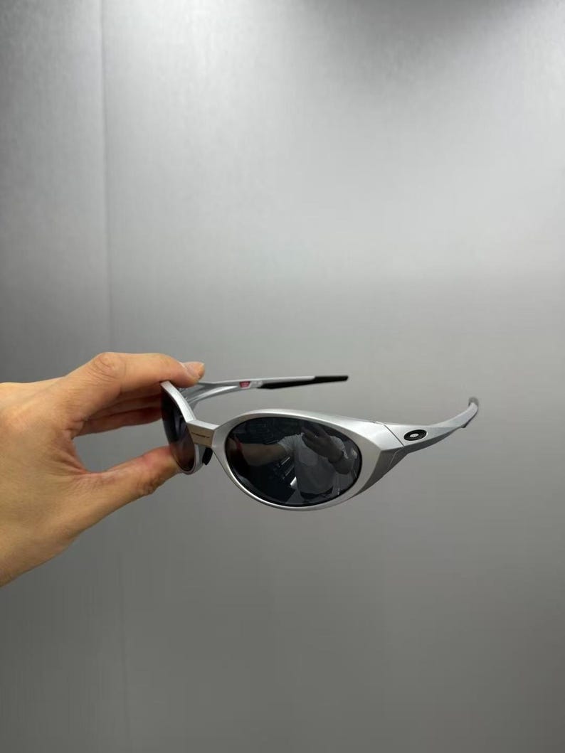 Pode incluir: &Oacute;culos de sol prateados com lentes escuras. Os &oacute;culos t&ecirc;m um design moderno com hastes curvas e uma forma de lente arredondada. As lentes escuras protegem do sol.