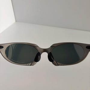 May include: Gray sunglasses with dark lenses. The sunglasses have a sleek, modern design with a metal frame and a black nose bridge. The lenses are oval-shaped and appear to be polarized.