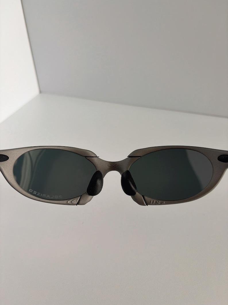 May include: Gray sunglasses with dark lenses. The sunglasses have a sleek, modern design with a metal frame and a black nose bridge. The lenses are oval-shaped and appear to be polarized.