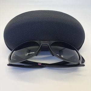 May include: Black sunglasses with a matching black case. The sunglasses have a sleek, modern design with dark lenses and a black frame. The case is oval-shaped and has a textured surface. The sunglasses are labeled "USA".