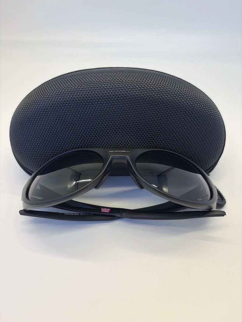 May include: Black sunglasses with a matching black case. The sunglasses have a sleek, modern design with dark lenses and a black frame. The case is oval-shaped and has a textured surface. The sunglasses are labeled "USA".