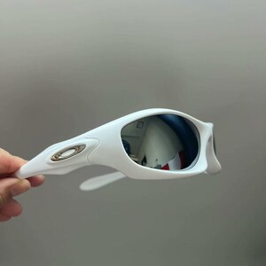 K&ouml;nnte beinhalten: Wei&szlig;e Sonnenbrille mit verspiegelten silbernen Gl&auml;sern. Die Sonnenbrille hat einen geschwungenen Rahmen mit einem metallischen Logo am B&uuml;gel. Die Gl&auml;ser reflektieren die Umgebung.