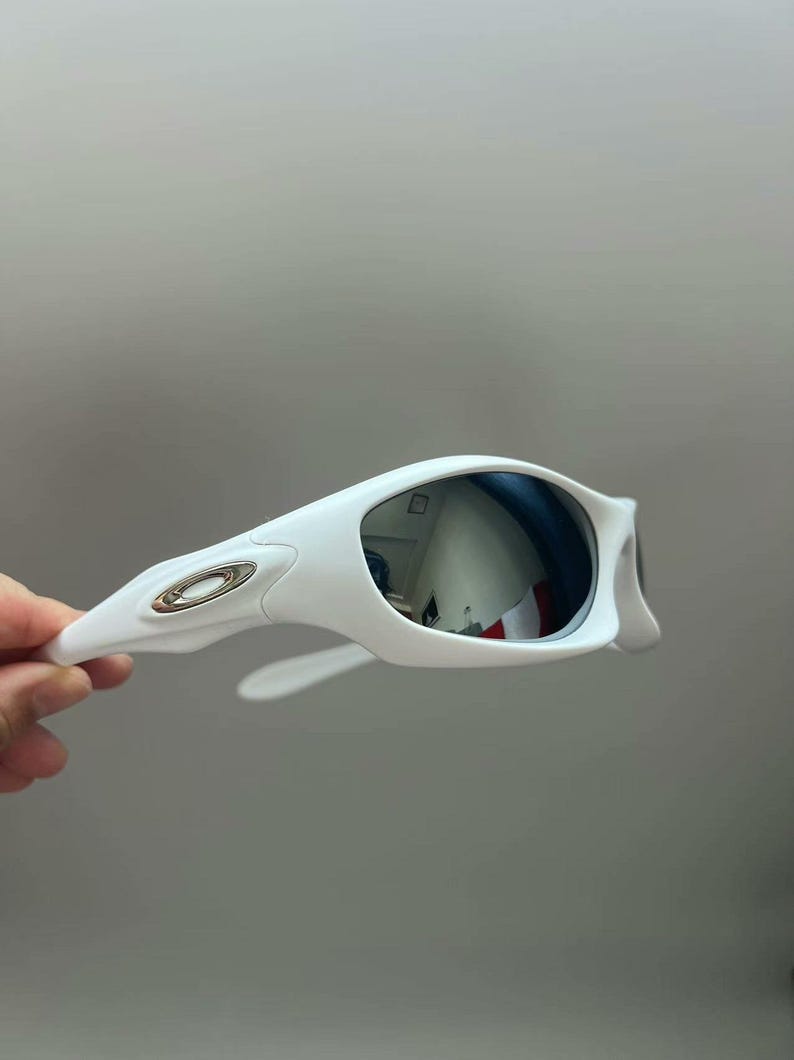 K&ouml;nnte beinhalten: Wei&szlig;e Sonnenbrille mit verspiegelten silbernen Gl&auml;sern. Die Sonnenbrille hat einen geschwungenen Rahmen mit einem metallischen Logo am B&uuml;gel. Die Gl&auml;ser reflektieren die Umgebung.