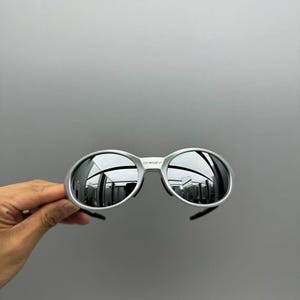 May include: Silver-framed sunglasses with mirrored lenses. The sunglasses have a rounded shape and a sleek, modern design. The lenses reflect the surrounding environment. The brand name "Oakley" is visible on the frame.