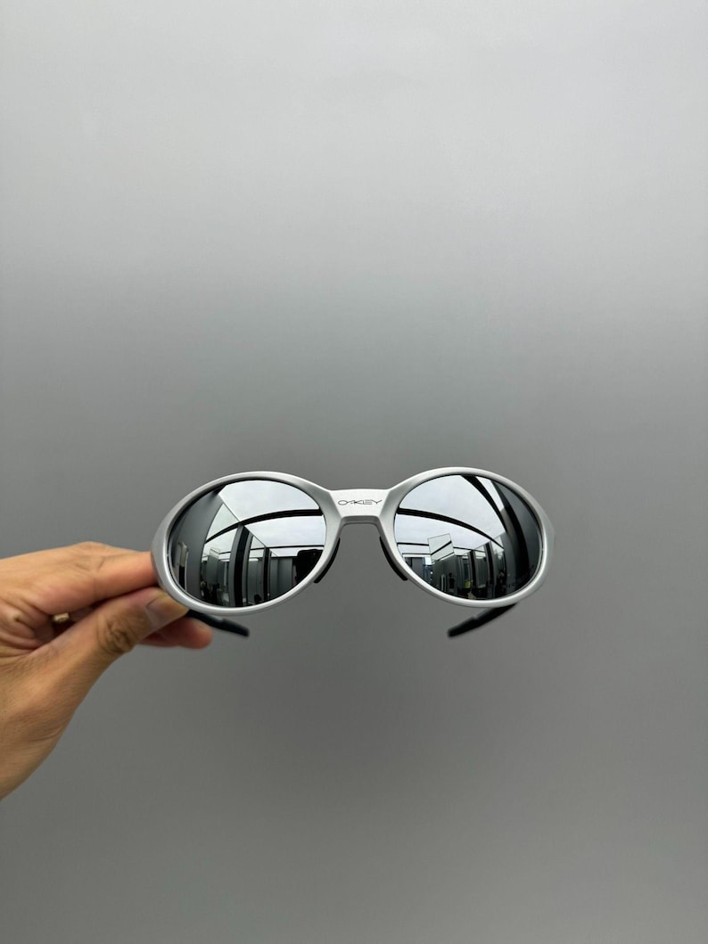 Pu&ograve; includere: Occhiali da sole con montatura argentata e lenti a specchio. Gli occhiali hanno una forma arrotondata e un design elegante. Le lenti riflettono l'ambiente circostante. Il marchio "Oakley" &egrave; visibile sulla montatura.