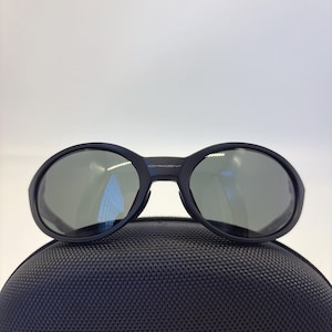 May include: Black sunglasses with oval lenses and a dark frame. The sunglasses are resting on a black, textured case. The Oakley logo is visible on the frame. The lenses have a reflective quality.