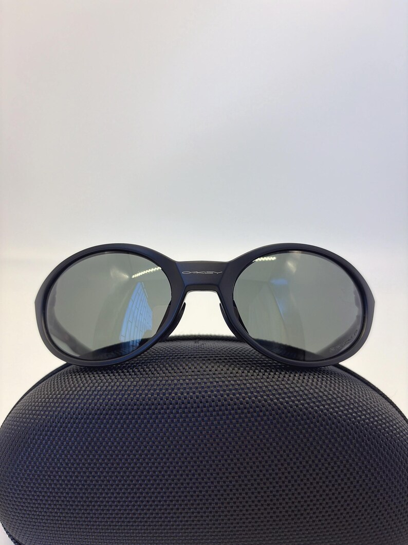 May include: Black sunglasses with oval lenses and a dark frame. The sunglasses are resting on a black, textured case. The Oakley logo is visible on the frame. The lenses have a reflective quality.