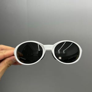 K&ouml;nnte beinhalten: Wei&szlig;e ovale Sonnenbrille mit dunklen Gl&auml;sern. Die Sonnenbrille hat einen dicken wei&szlig;en Rahmen und einen Steg, der die beiden Gl&auml;ser verbindet. Die Gl&auml;ser sind dunkel und reflektieren die Umgebung. Die Sonnenbrille wird von einer Hand gehalten.