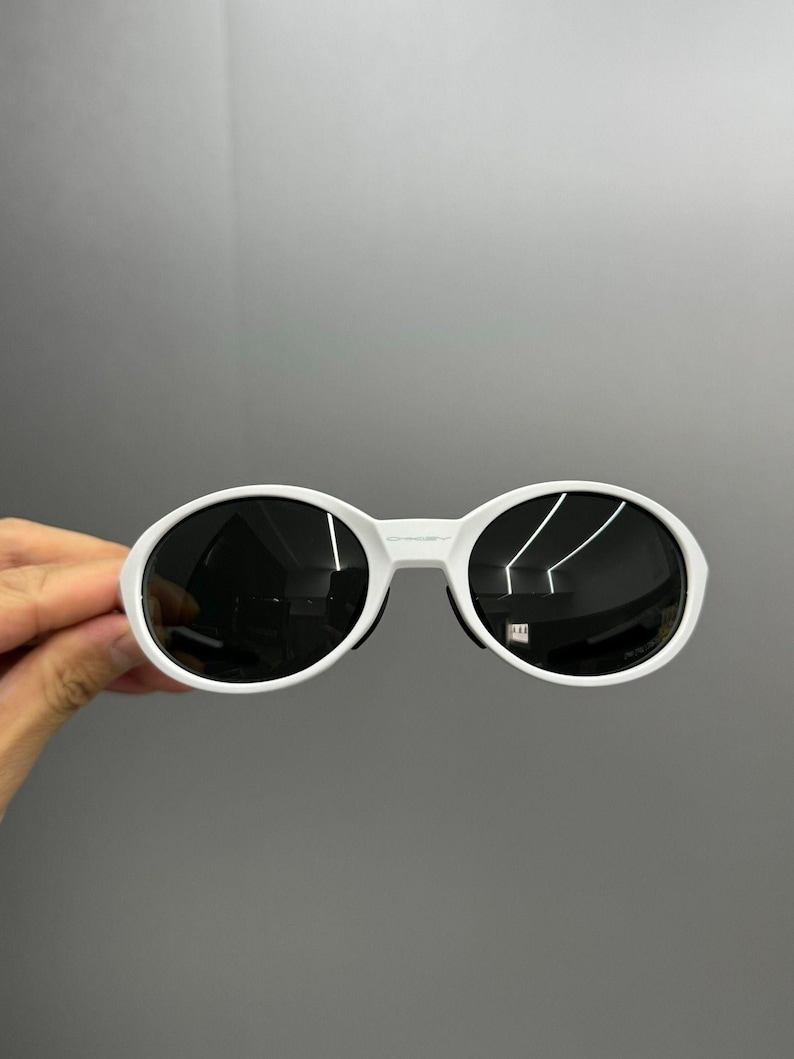 K&ouml;nnte beinhalten: Wei&szlig;e ovale Sonnenbrille mit dunklen Gl&auml;sern. Die Sonnenbrille hat einen dicken wei&szlig;en Rahmen und einen Steg, der die beiden Gl&auml;ser verbindet. Die Gl&auml;ser sind dunkel und reflektieren die Umgebung. Die Sonnenbrille wird von einer Hand gehalten.