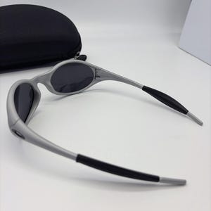 Puede incluir: Gafas de sol grises con lentes oscuras y puntas de patillas negras sobre una superficie blanca. Un estuche negro semicircular para gafas en el fondo. Dise&ntilde;o deportivo y aerodin&aacute;mico.