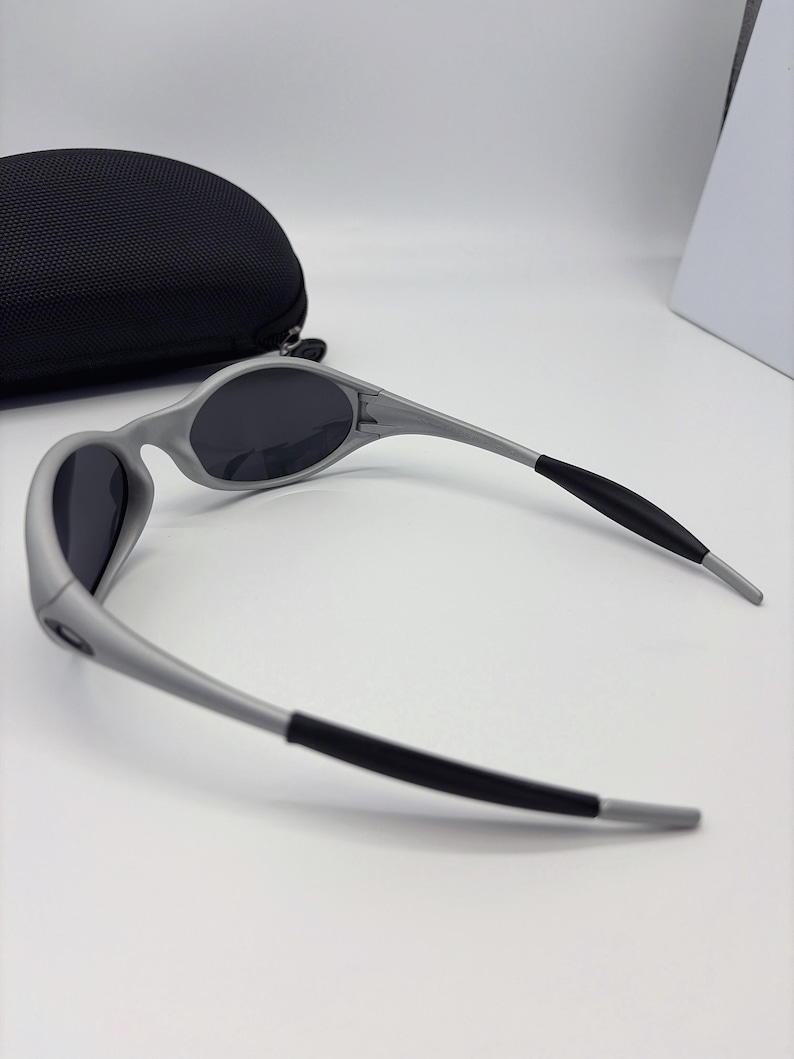 Puede incluir: Gafas de sol grises con lentes oscuras y puntas de patillas negras sobre una superficie blanca. Un estuche negro semicircular para gafas en el fondo. Dise&ntilde;o deportivo y aerodin&aacute;mico.