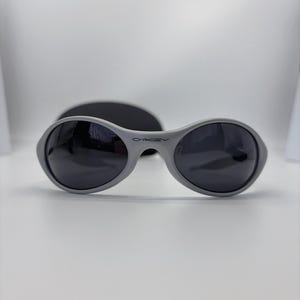 Puede incluir: Gafas de sol grises con lentes oscuras y el nombre de la marca "Oakley" impreso en la montura. Las gafas de sol tienen forma redondeada y est&aacute;n sobre un fondo blanco. Se ve un estuche negro detr&aacute;s de las gafas.