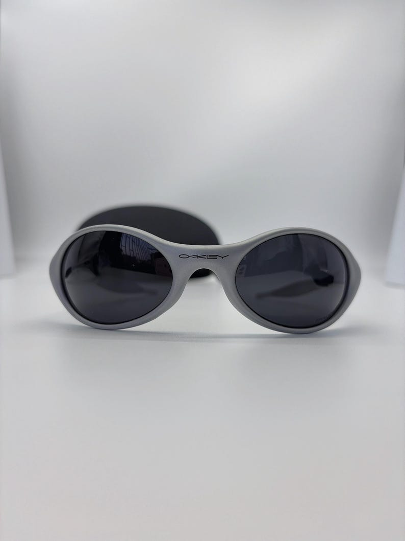 Puede incluir: Gafas de sol grises con lentes oscuras y el nombre de la marca "Oakley" impreso en la montura. Las gafas de sol tienen forma redondeada y est&aacute;n sobre un fondo blanco. Se ve un estuche negro detr&aacute;s de las gafas.