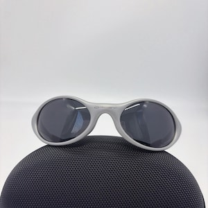 Puede incluir: Gafas de sol Oakley grises con lentes oscuras sobre un estuche negro texturizado. Las gafas de sol tienen un dise&ntilde;o de montura redondeada con el logotipo de Oakley en el puente. El estuche tiene cremallera y forma redondeada.