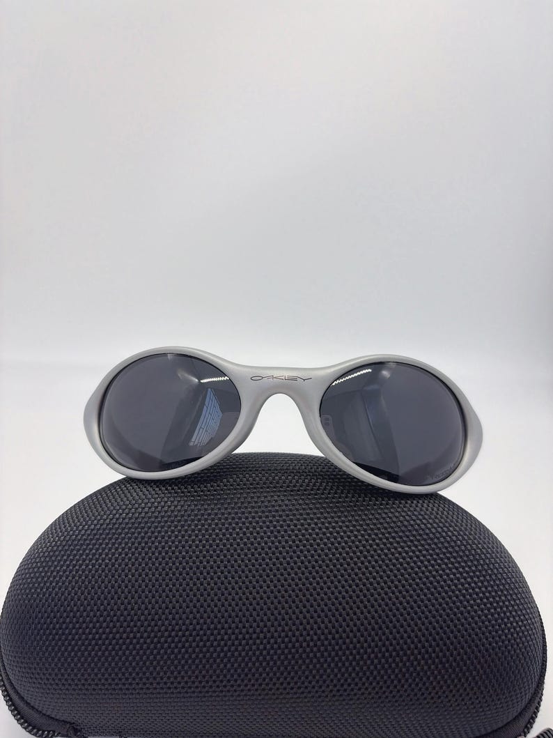 Puede incluir: Gafas de sol Oakley grises con lentes oscuras sobre un estuche negro texturizado. Las gafas de sol tienen un dise&ntilde;o de montura redondeada con el logotipo de Oakley en el puente. El estuche tiene cremallera y forma redondeada.