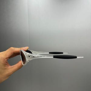 May include: Clear-framed sunglasses with dark lenses and black temples. The sunglasses are held up, showcasing the design and shape. The frame has a sleek, modern design, suitable for outdoor activities.